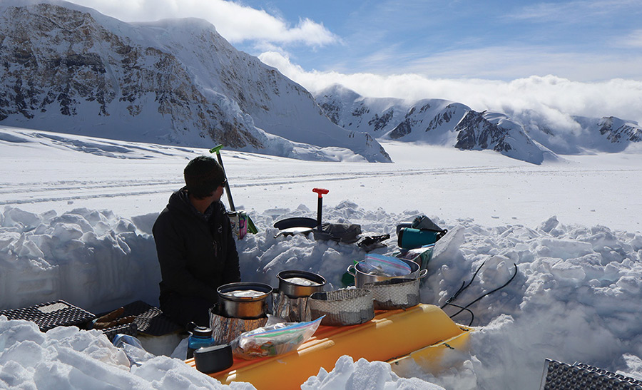 A person sits in a snow trench camp set up with cooking gear, looking out across a vast, snow-covered mountain landscape.