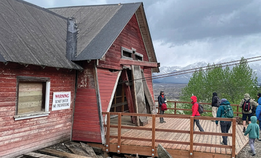 Visitors gather on a wooden deck outside a historic red wooden building with a warning sign, looking out toward distant mountains.