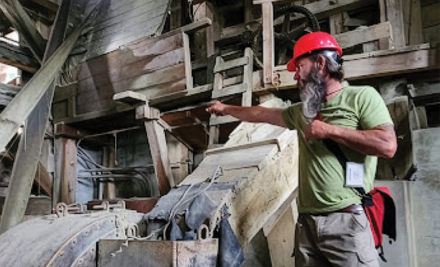 A man wearing a red hard hat points toward historic wooden mining equipment inside an industrial structure.