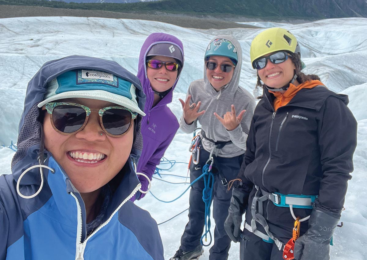 Four smiling hikers in helmets, climbing gear, and sunglasses pose for a group selfie while trekking on a glacier.
