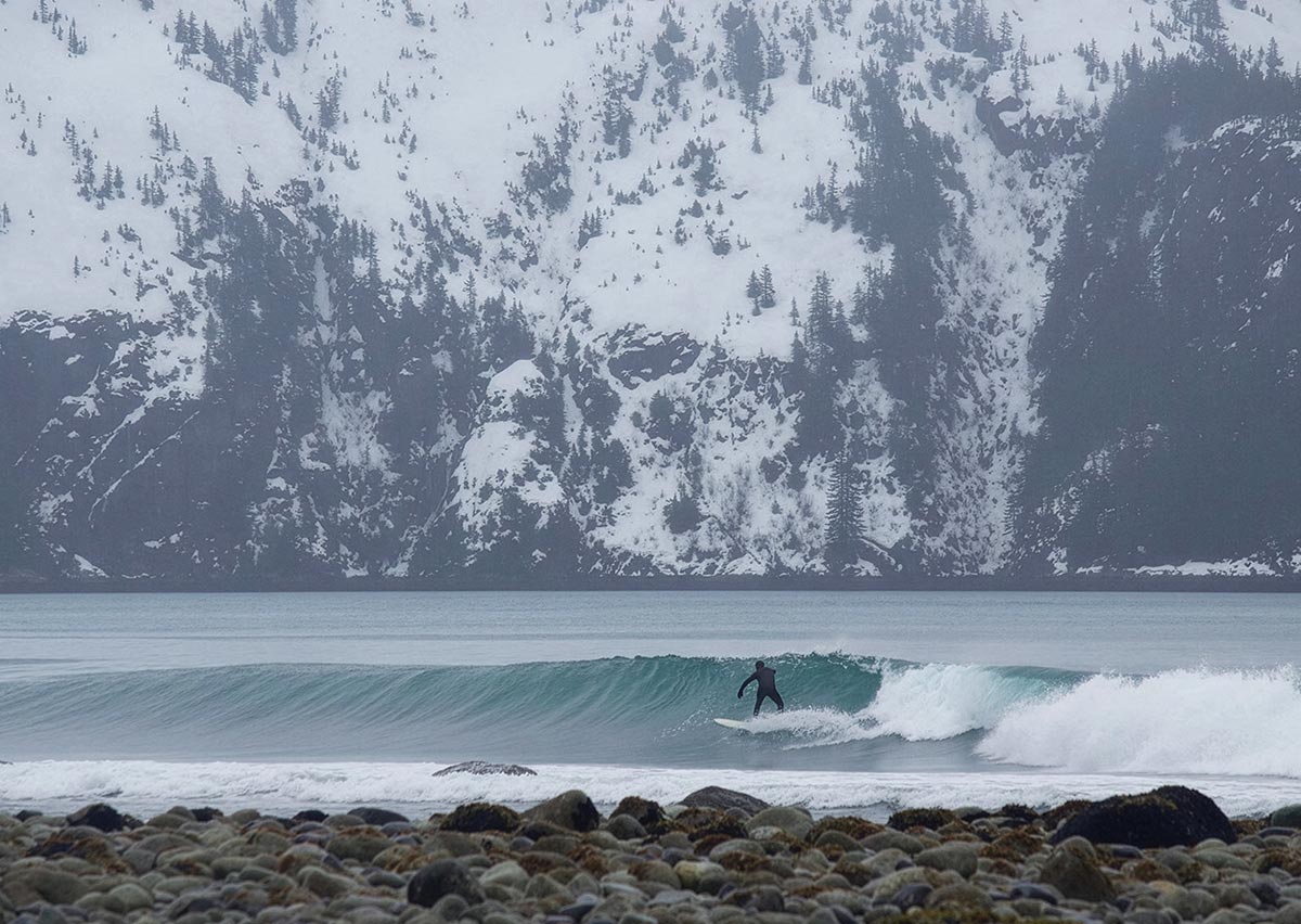 A surfer in a wetsuit rides an ocean wave along a rocky shore, with steep, snow-covered mountains in the background.