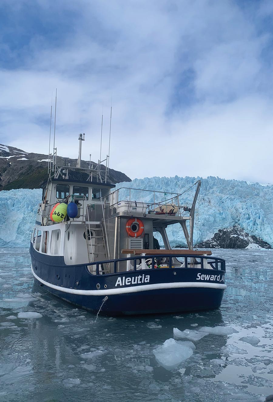 A boat named Aleutia from Seward, AK navigates through ice-filled waters in front of a massive blue glacier.