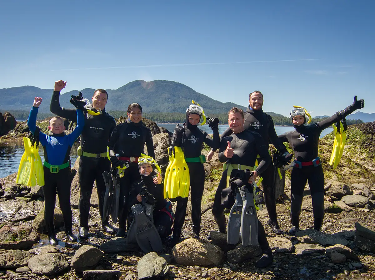A group of smiling scuba divers and snorkelers in wetsuits hold up their fins and pose along a rocky shoreline with water and mountains in the background.