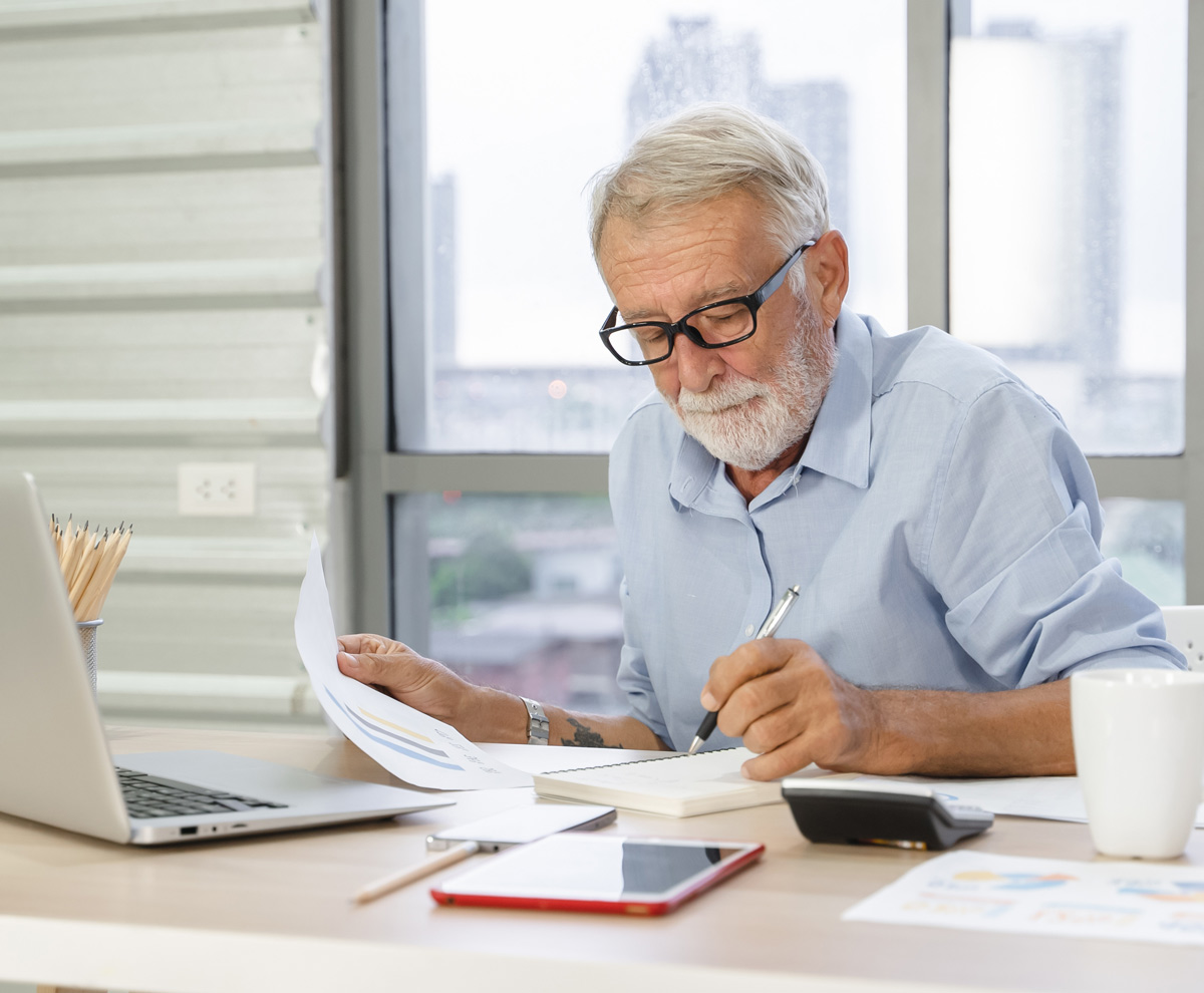 Senior man with a white beard and glasses focused on writing in a notebook while reviewing paper documents at a desk.
