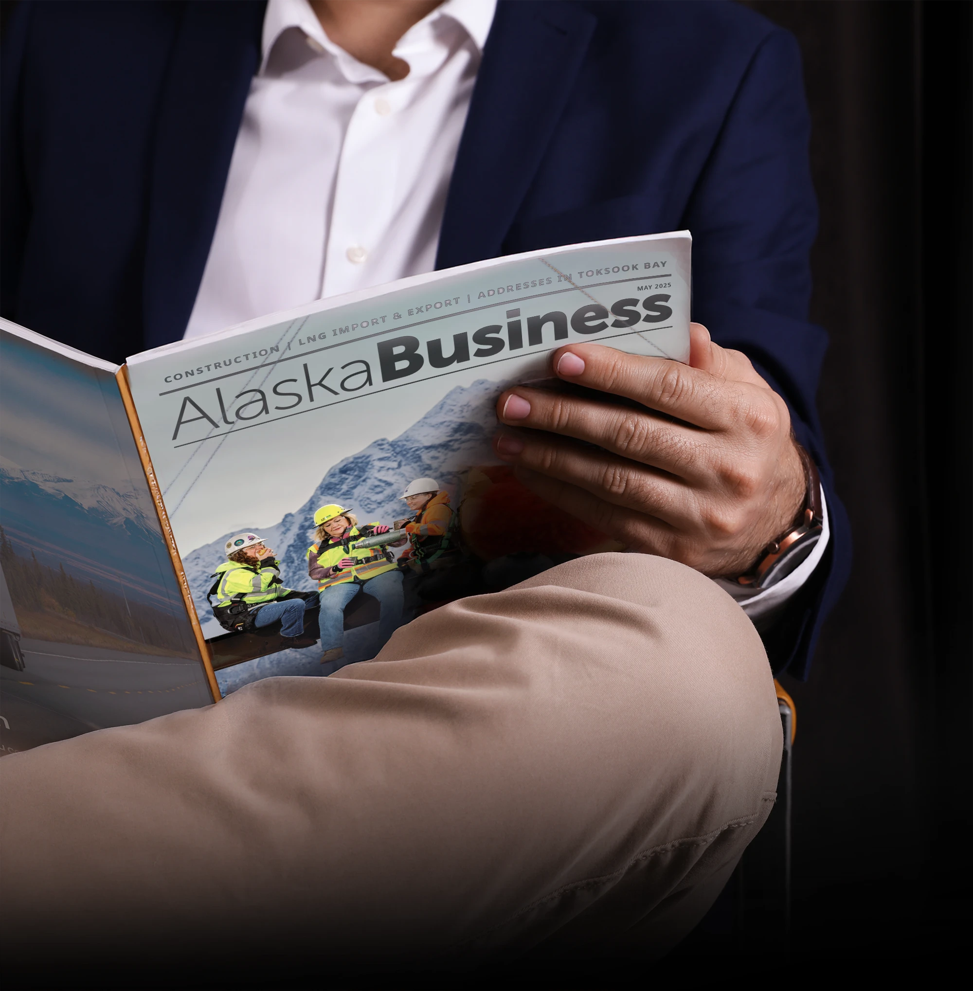 Person in a suit holding and reading a copy of Alaska Business magazine with a cover featuring construction workers in safety gear.