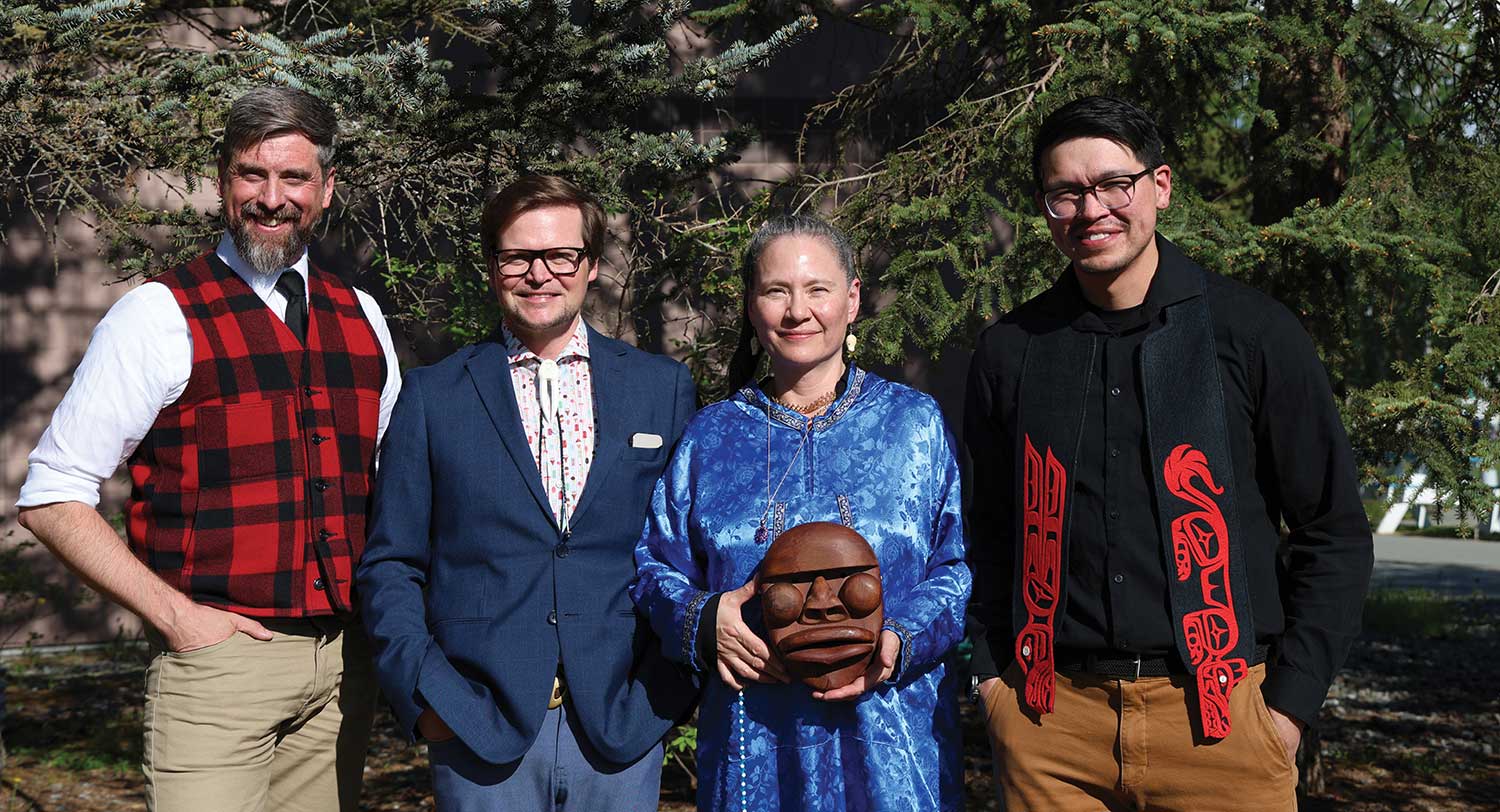 Four people standing outdoors together, including a woman in blue holding a carved mask.