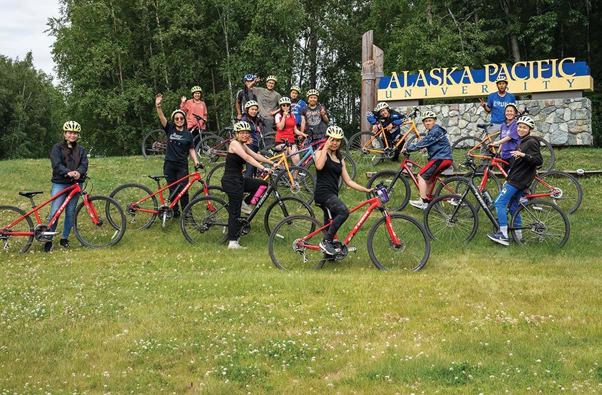 Group of cyclists with bicycles posing on a grassy hill in front of Alaska Pacific University sign.