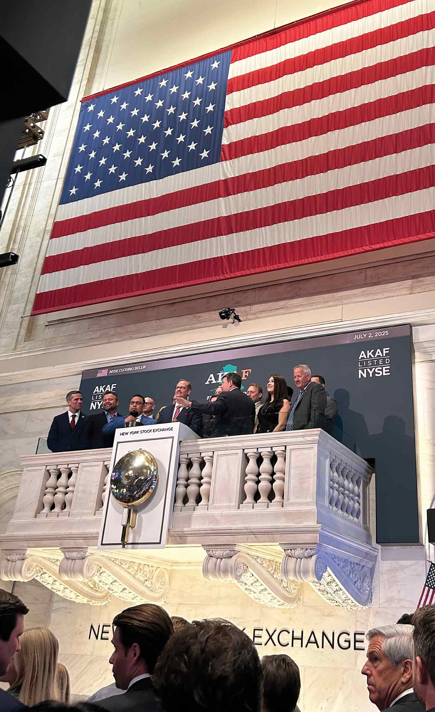 Group standing on the New York Stock Exchange balcony under a large American flag.
