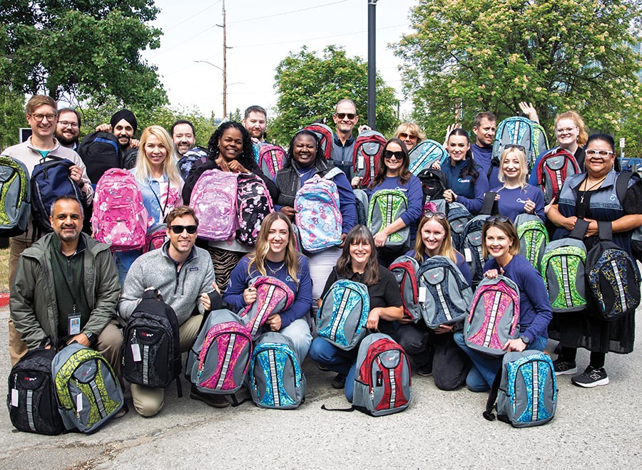 A large group of volunteers posing outdoors while holding colorful school backpacks.