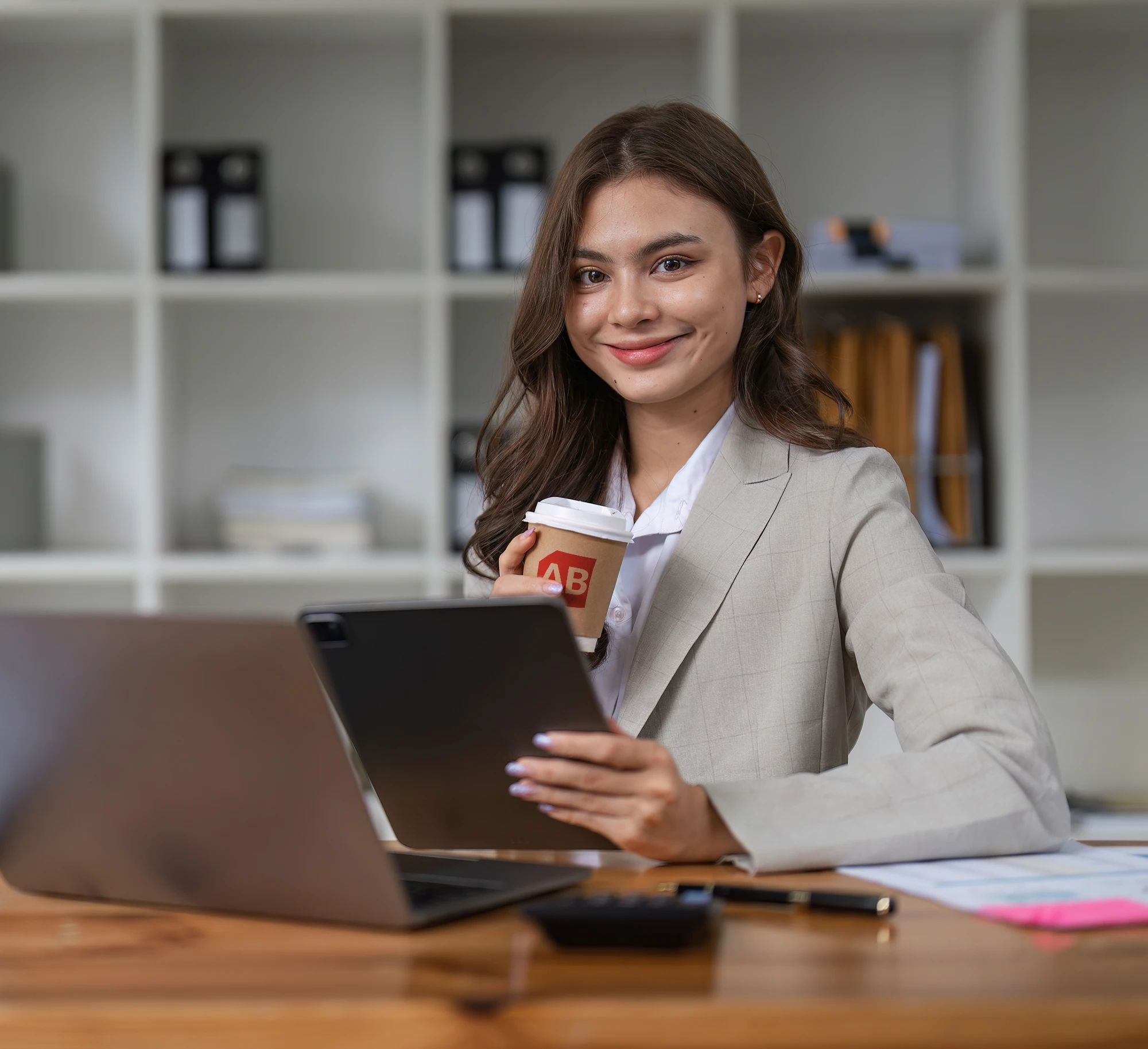 A business professional holds a tablet and an Alaska Business-branded coffee cup while working at a laptop in an office.