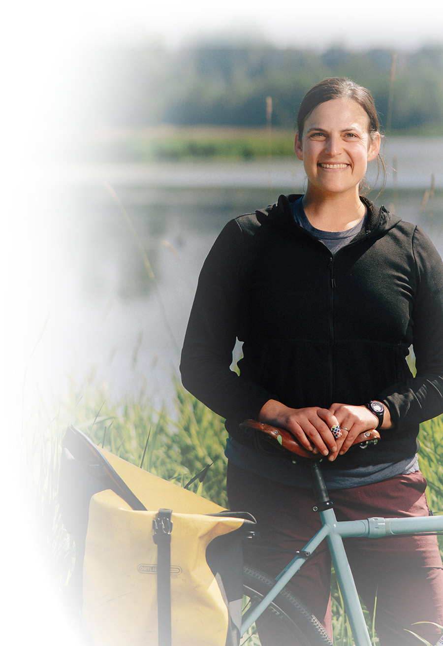 A smiling woman standing outdoors next to a bicycle equipped with a yellow pannier bag.