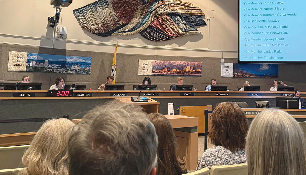 Audience members watching a city council meeting panel seated in front of voting displays.